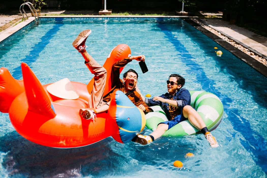 Two men sitting on pool floating toys with all of their clothes on in the pool.