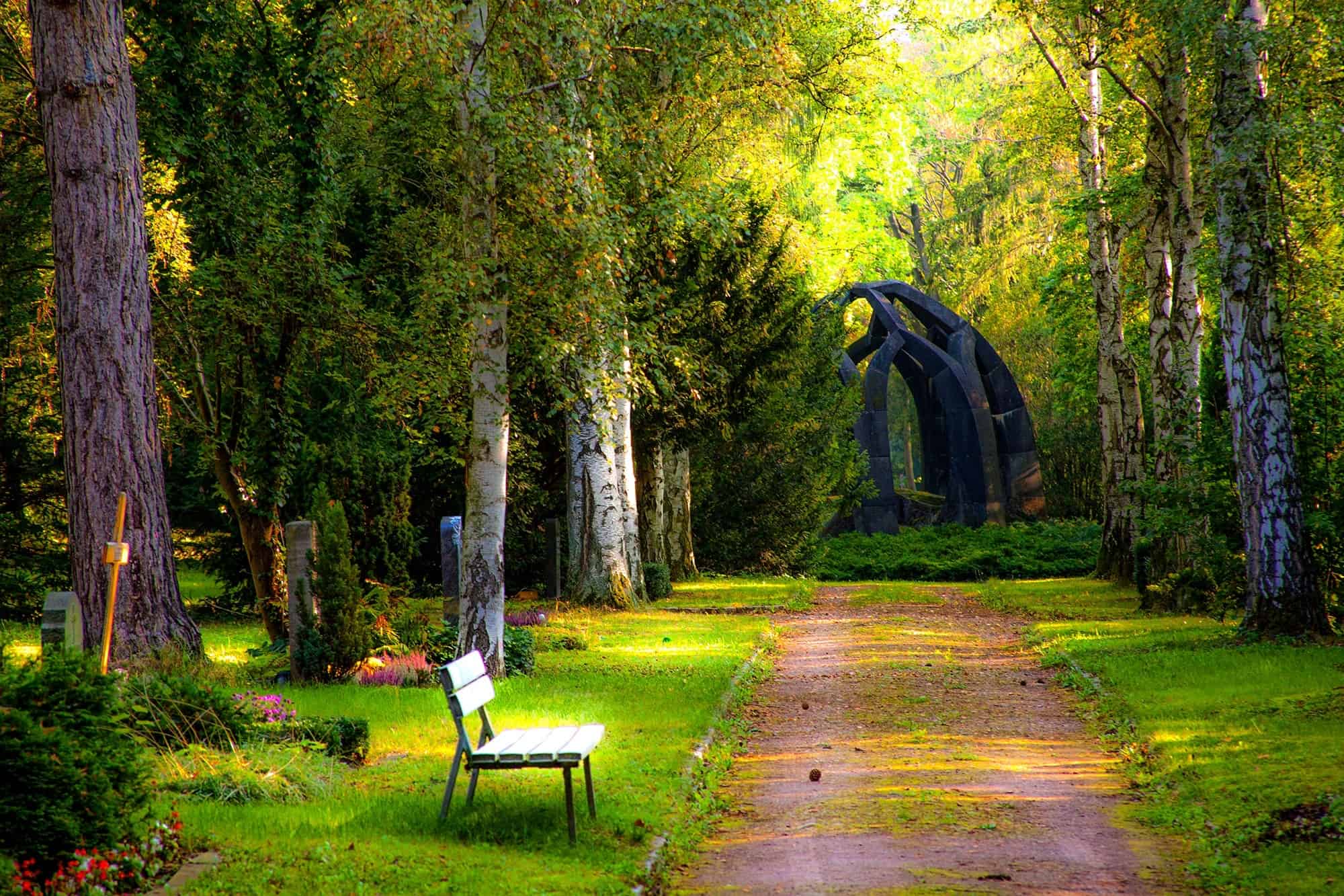 A park with green grass and trees and a bench in the foreground and a large black statue in the background.