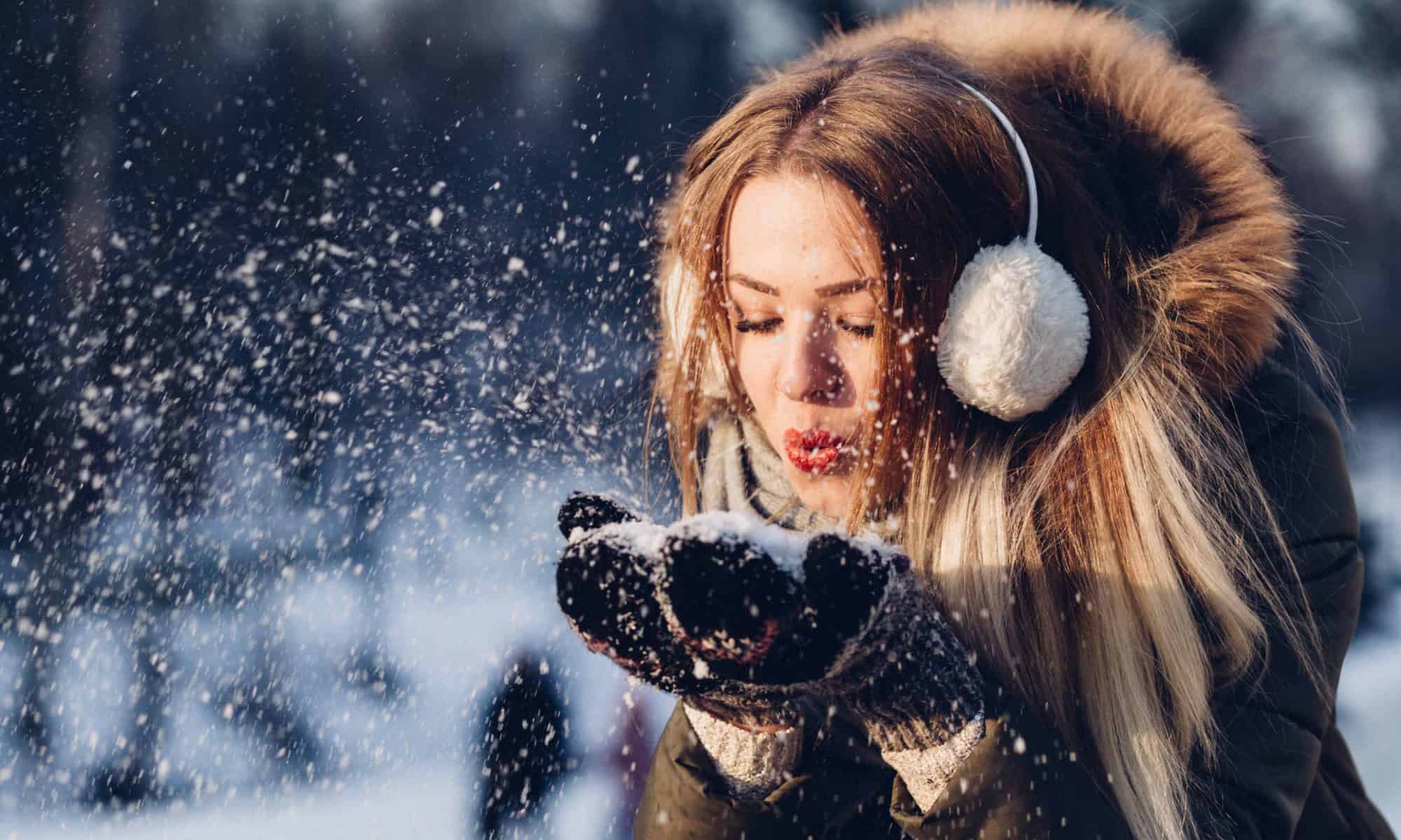 A woman with a large jacket and earmuffs blowing snow out of her hands.