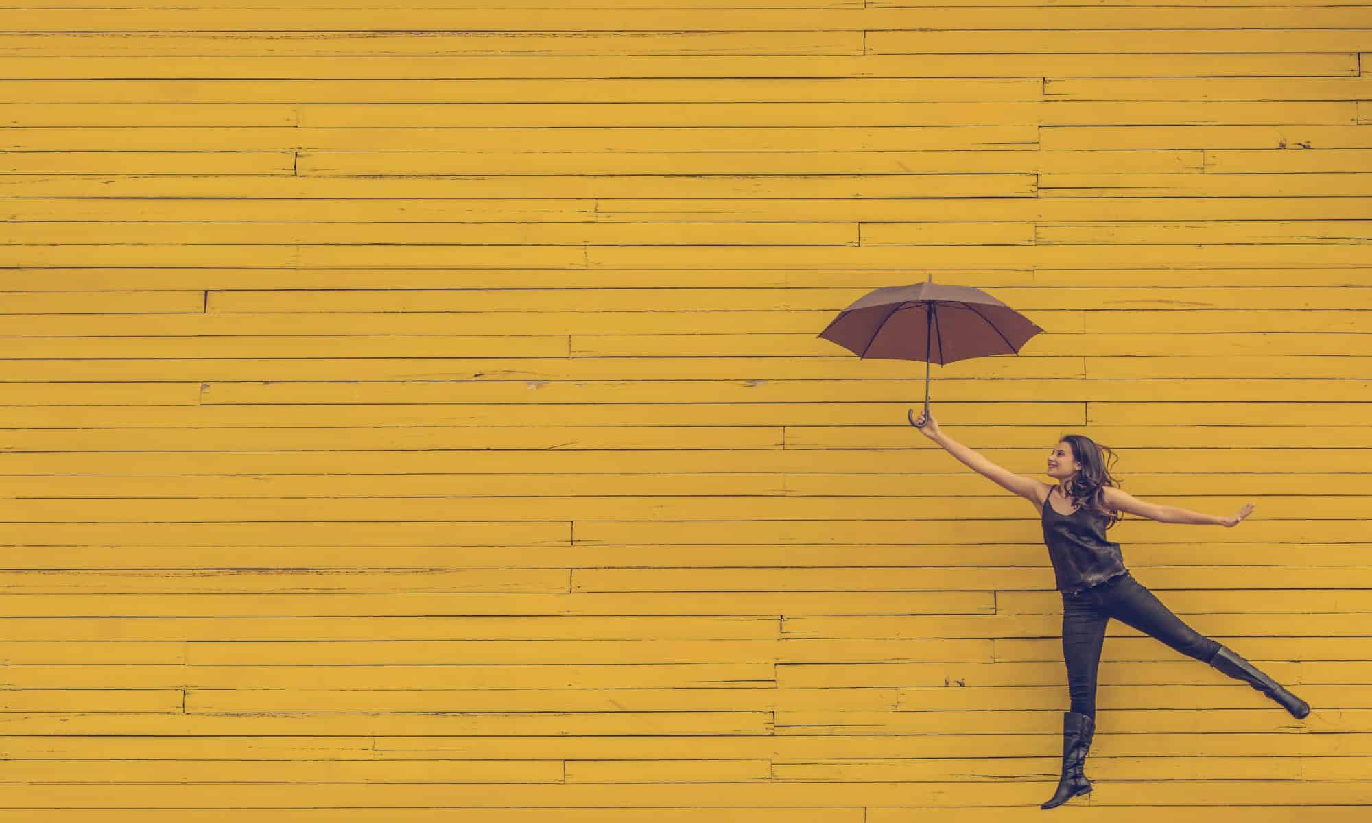 Young girl holding purple umbrella and jumping in front of mustard-coloured wall.