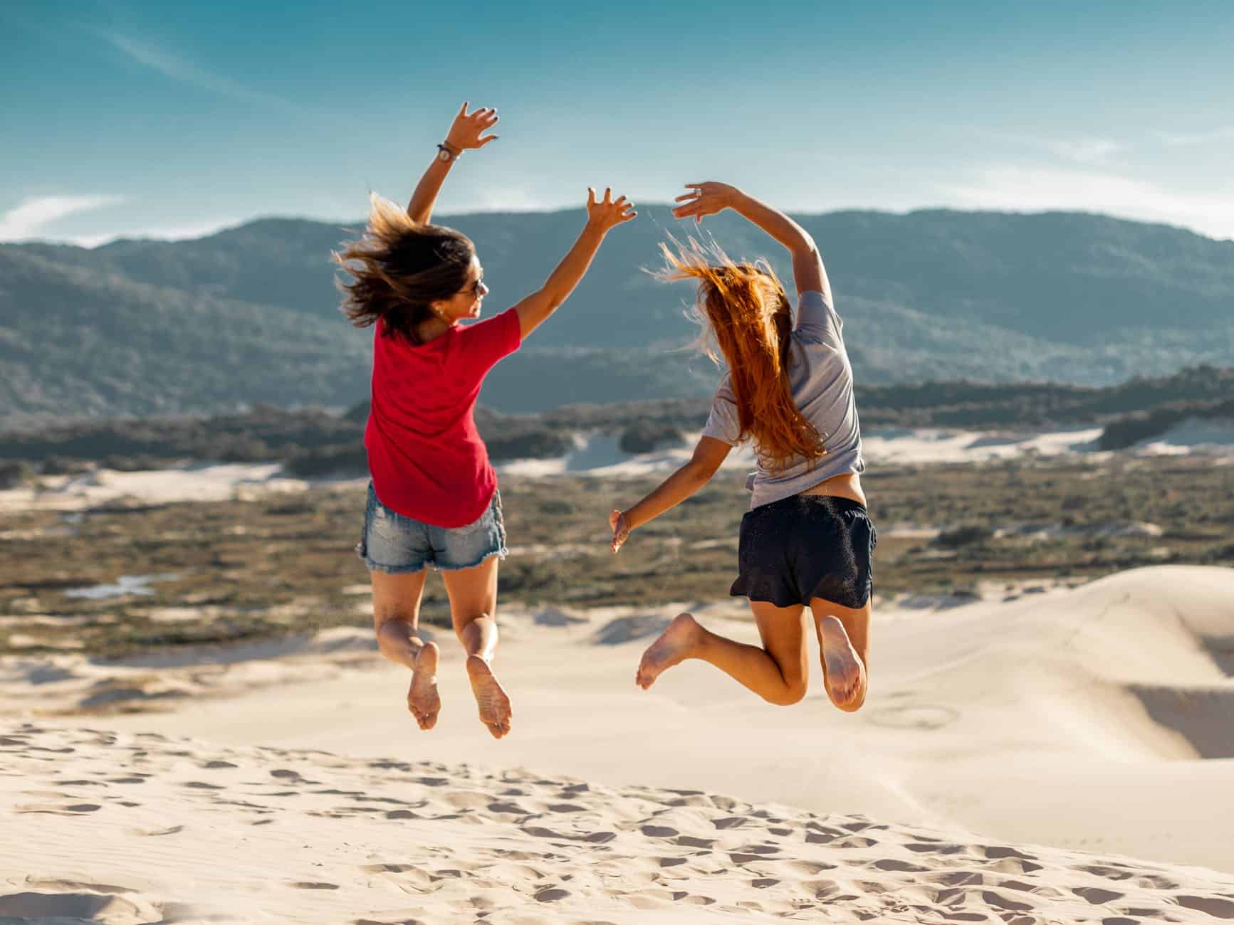 Two young people jumping on the sand with mountains in the background
