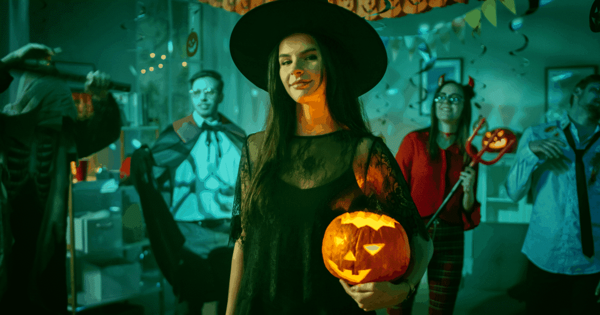 Young woman holding a carved pumpkin in a witch costume with other people dressed up behind her.