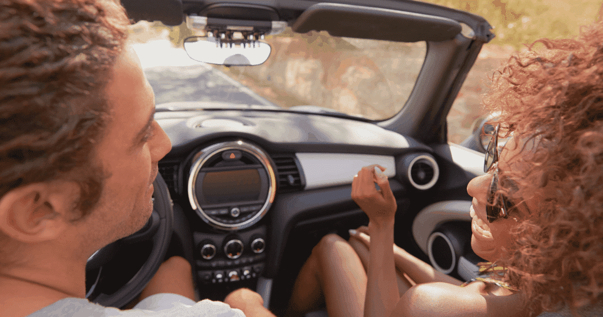 A young man and woman looking at each other while the man drives a convertible car.